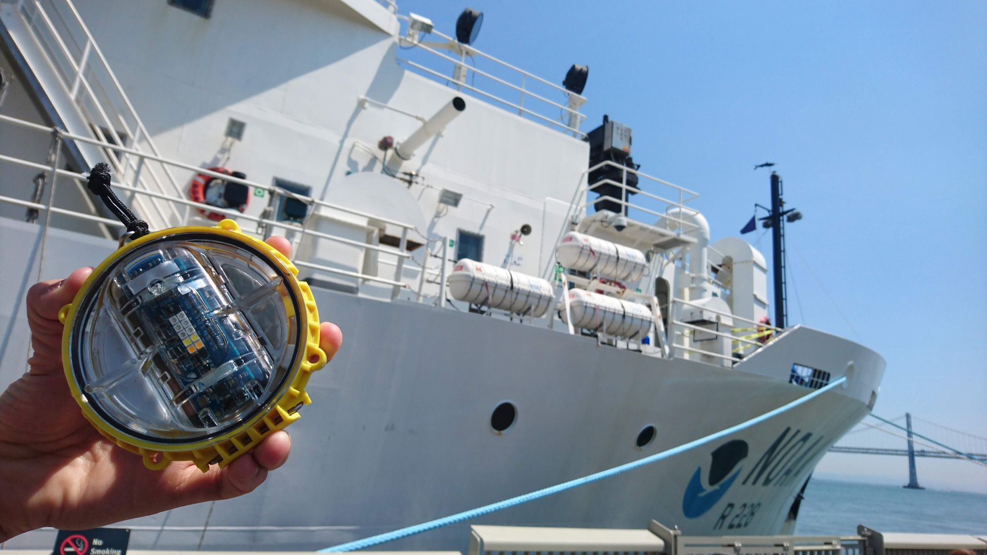 A photo of a hand holding the Pisces light, a round disc encased in yellow plastic, with a ship and sea in the background