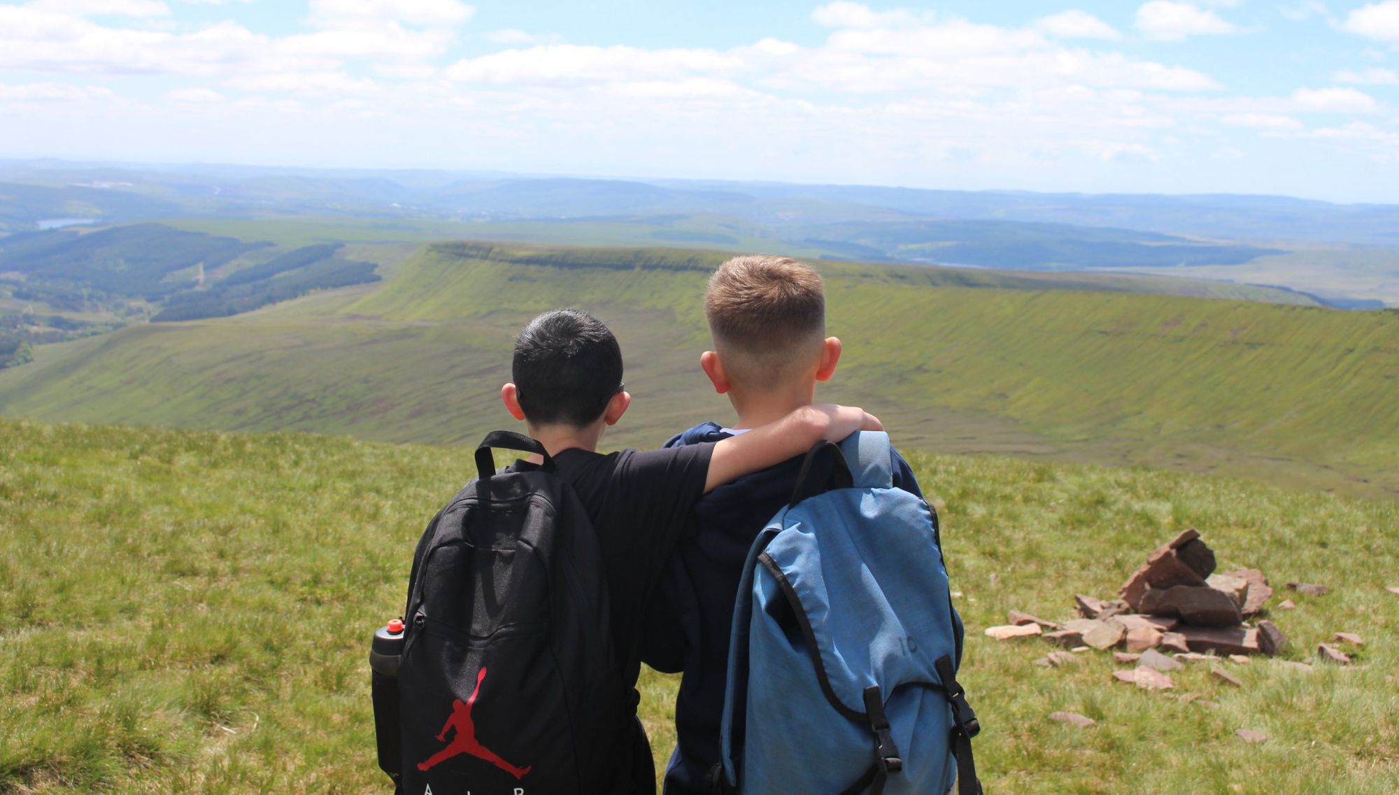 Two children in backpacks stand looking out over a moorland view, with their backs to the camera